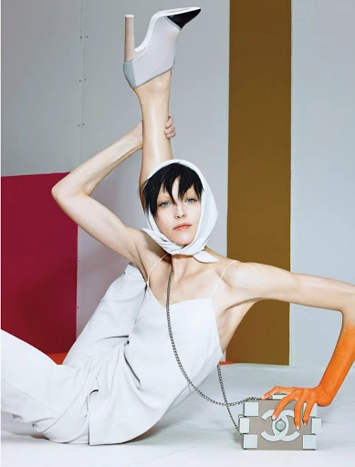 Woman in white outfit poses with high-heeled shoe and fashion purse in front of colorful graphic background in a studio setting.