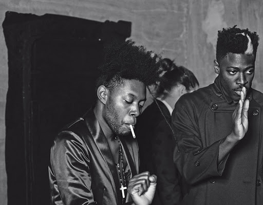 Black and white image featuring two stylish young men, one smoking a cigarette and wearing a cross necklace, in an indoor setting.
