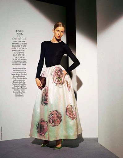 Fashion editorial shot of a model wearing a black long-sleeved top and a floral skirt, holding a clutch in a studio setting.