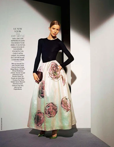 Fashion editorial shot of a model wearing a black long-sleeved top and a floral skirt, holding a clutch in a studio setting.