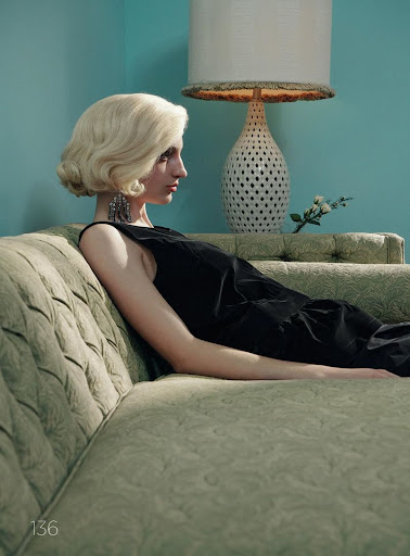 Profile of a woman with blonde wavy hair relaxing on a green vintage couch with a table lamp and turquoise wall in the background.