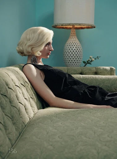 Profile of a woman with blonde wavy hair relaxing on a green vintage couch with a table lamp and turquoise wall in the background.
