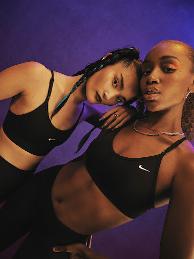 Two women in black sports bras and leggings pose against a purple backdrop, showcasing fitness apparel in a studio setting.