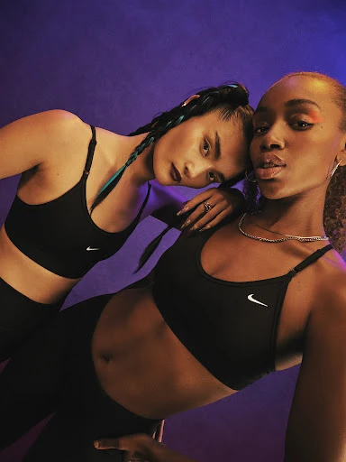 Two women in black sports bras and leggings pose against a purple backdrop, showcasing fitness apparel in a studio setting.