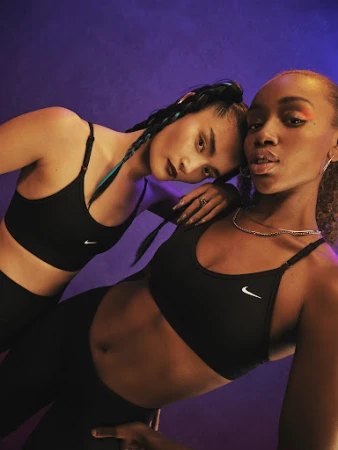 Two women in black sports bras and leggings pose against a purple backdrop, showcasing fitness apparel in a studio setting.