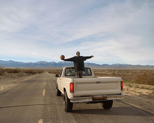 Man standing in the back of a truck holding a basketball on a desert highway with mountains in the distance under a partly cloudy sky.