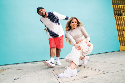 Two people, a man and woman, pose in front of a bright turquoise wall. The man is standing while the woman is crouching and smiling.