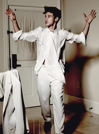 A young adult male in a white suit stands with his arms raised in an indoor setting with white walls and a suit jacket hanging nearby.
