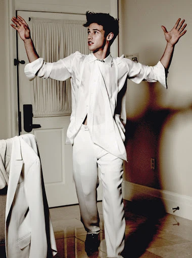 A young adult male in a white suit stands with his arms raised in an indoor setting with white walls and a suit jacket hanging nearby.