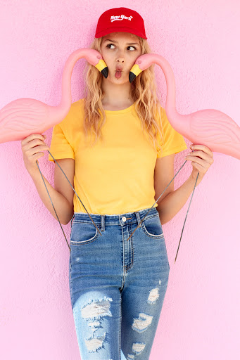 Blonde woman in a yellow t-shirt and a red cap is holding two flamingo props against a light pink wall in an eye-level shot.