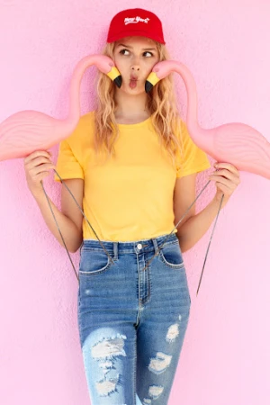 Blonde woman in a yellow t-shirt and a red cap is holding two flamingo props against a light pink wall in an eye-level shot.