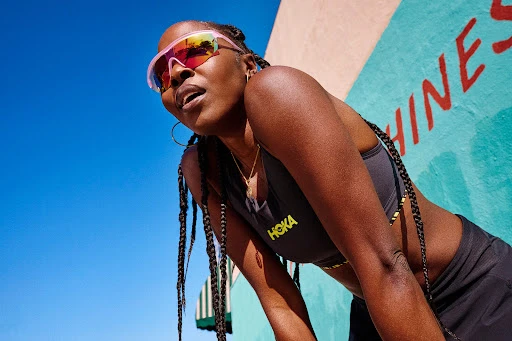 Athletic woman in sunglasses with braided hair, wearing a sports bra and skirt, stands near a colorful wall against a blue sky.