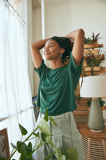 Smiling woman with braided hair standing near a window in a bright room, wearing a green t-shirt and light-colored bottoms.