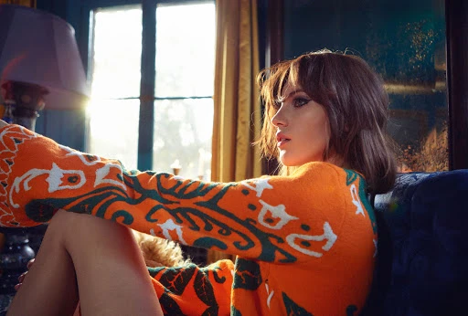Brunette woman with bangs sits on blue velvet couch wearing an orange patterned sweater in interior lighting near a window.
