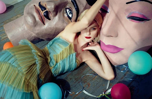 Blonde woman in blue and gold dress lying on floor with oversized mask props and colorful balloons for a surreal fashion shot.