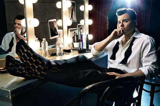 A man with dark hair and a black bow tie sitting at a lighted vanity table, legs crossed and feet up, wearing patterned socks.