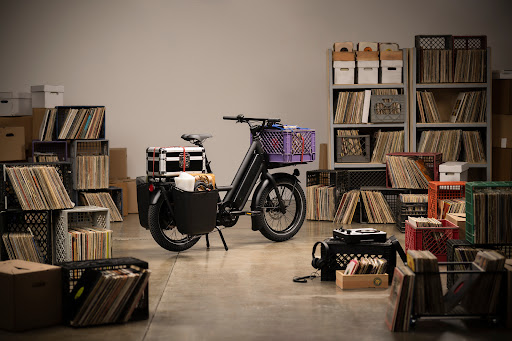 Black moped parked indoors with vinyl records in milk crates and boxes, creating a vintage music storage and transportation setup.