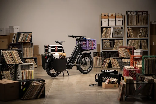 Black moped parked indoors with vinyl records in milk crates and boxes, creating a vintage music storage and transportation setup.