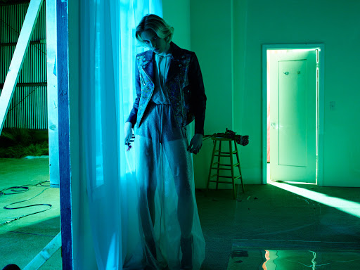 Adult with blond hair poses in sheer dress and leather jacket next to curtains in a green-lit room with a closed door.