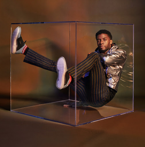 Portrait of a man wearing a metallic jacket and striped pants, sitting inside a transparent cube against a gradient brown background.