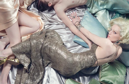 Fashion editorial with woman in gold sequin dress lying on satin sheets, accompanied by two other people partially visible.