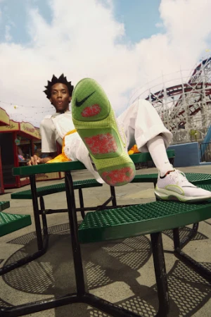 Low-angle shot of an adult wearing white sneakers with a lime green sole, sitting on a green metal picnic table at an amusement park.