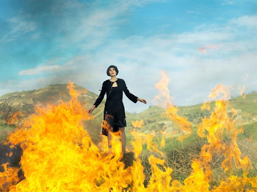 Woman stands in a black dress surrounded by flames against a background of grassy hills and a blue, cloudy sky in an outdoor setting.