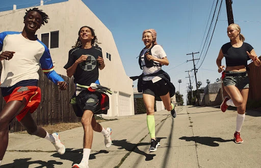 Four adult runners - three women and one man - are jogging on an urban street during the daytime. They are dressed in athletic gear.