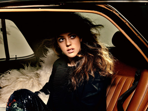 Young woman with wavy hair in car. She's wearing a black jacket and shiny pants. The car has tan leather seats.