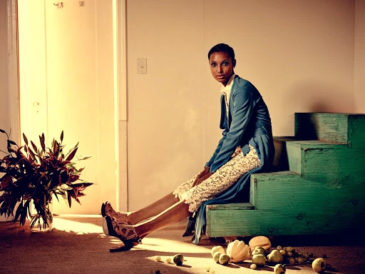 Model seated on teal wooden steps wearing a blue coat and patterned skirt, next to flowers and scattered fruit on the floor.