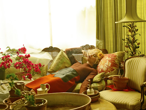 A woman with blond hair reclines on a vintage couch, smoking a cigarette. She is surrounded by potted plants and pillows in a warm-toned indoor setting.