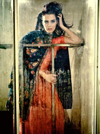 Fashion shot of a woman with dark hair wearing a patterned cape and peach dress, leaning on a metal frame in a mottled setting.
