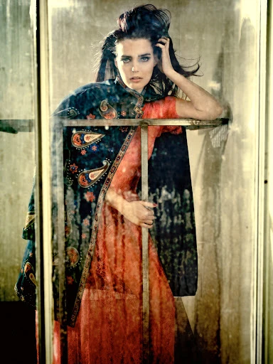 Fashion shot of a woman with dark hair wearing a patterned cape and peach dress, leaning on a metal frame in a mottled setting.