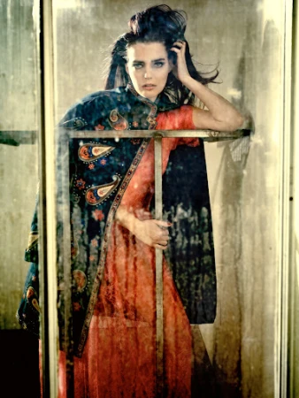 Fashion shot of a woman with dark hair wearing a patterned cape and peach dress, leaning on a metal frame in a mottled setting.