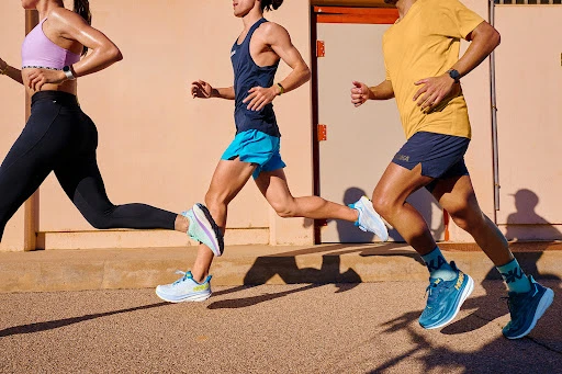Three athletes are running on a paved surface, wearing athletic clothing and running shoes, with an urban-style background. Focus on fitness and activity.