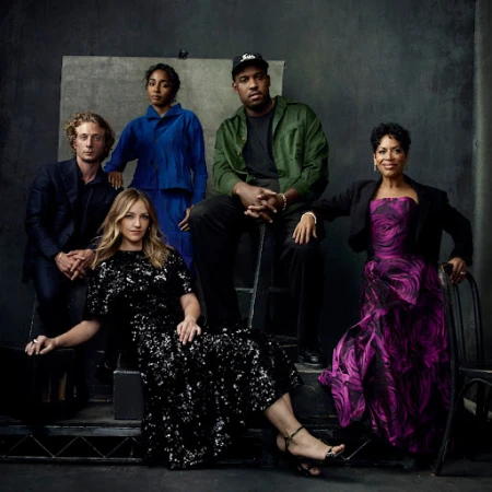 Group studio portrait of five adults, including one seated in a black sequin dress, and another wearing a formal pink gown, indoors.
