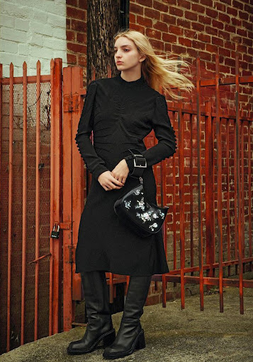 A blonde female model poses in a black dress and boots, holding a black purse, against a red brick wall and metal gate backdrop.