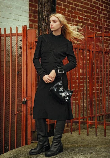 A blonde female model poses in a black dress and boots, holding a black purse, against a red brick wall and metal gate backdrop.