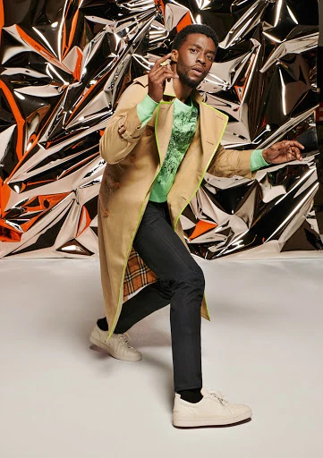 Celebrity posing in tan trench coat with lime green trim and light green sweater in front of reflective silver background in studio.
