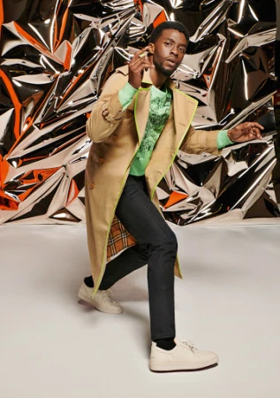 Celebrity posing in tan trench coat with lime green trim and light green sweater in front of reflective silver background in studio.