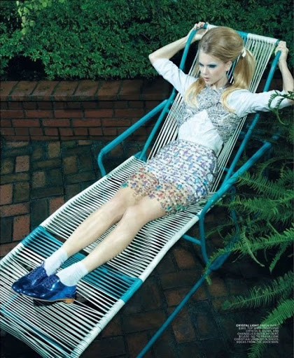 A young woman reclines on a turquoise lounge chair in a yard, wearing a shimmering silver top, floral skirt, socks, and patent blue shoes.