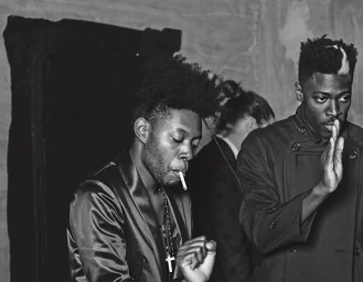 Black and white portrait of two Black men, one smoking with a cross necklace and another holding a hand up.