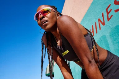 Black woman wearing pink reflective sports sunglasses and a dark athletic top against a blue and green wall