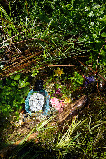 Blue perfume bottle nestled among green grass, yellow wildflowers, and a weathered log in a sun-drenched outdoor setting, nature.
