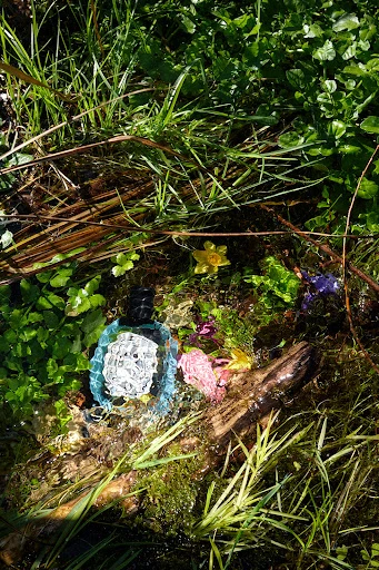 Blue perfume bottle nestled among green grass, yellow wildflowers, and a weathered log in a sun-drenched outdoor setting, nature.