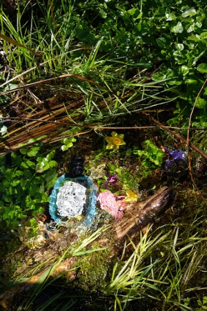 Blue perfume bottle nestled among green grass, yellow wildflowers, and a weathered log in a sun-drenched outdoor setting, nature.