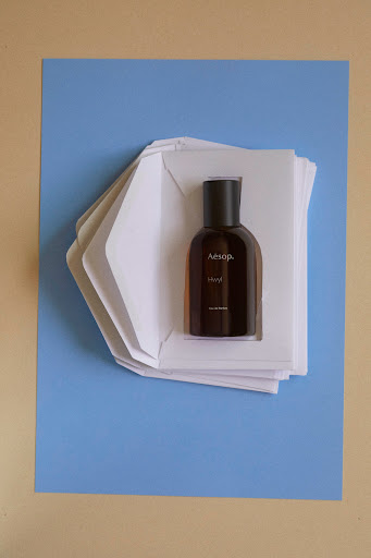 Amber glass Aesop perfume bottle sits among layered white envelopes on a pale blue surface against a tan background in studio lighting.