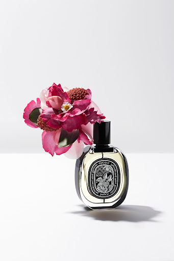 Perfume bottle with black cap next to pink and white flowers on a white surface and background in a product photography shot.