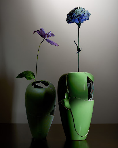 Still life of two green ceramic vases, one with a purple flower, the other with a blue carnation, set against a muted grey background.