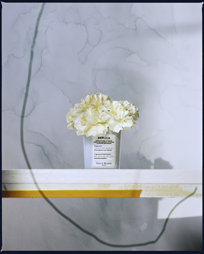 Still life of white carnations in a white Maison Margiela vase, sitting on a white shelf against a patterned light blue background.